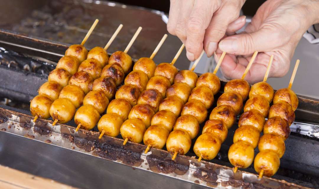 a griddle toasting mochi balls on sticks held by hands