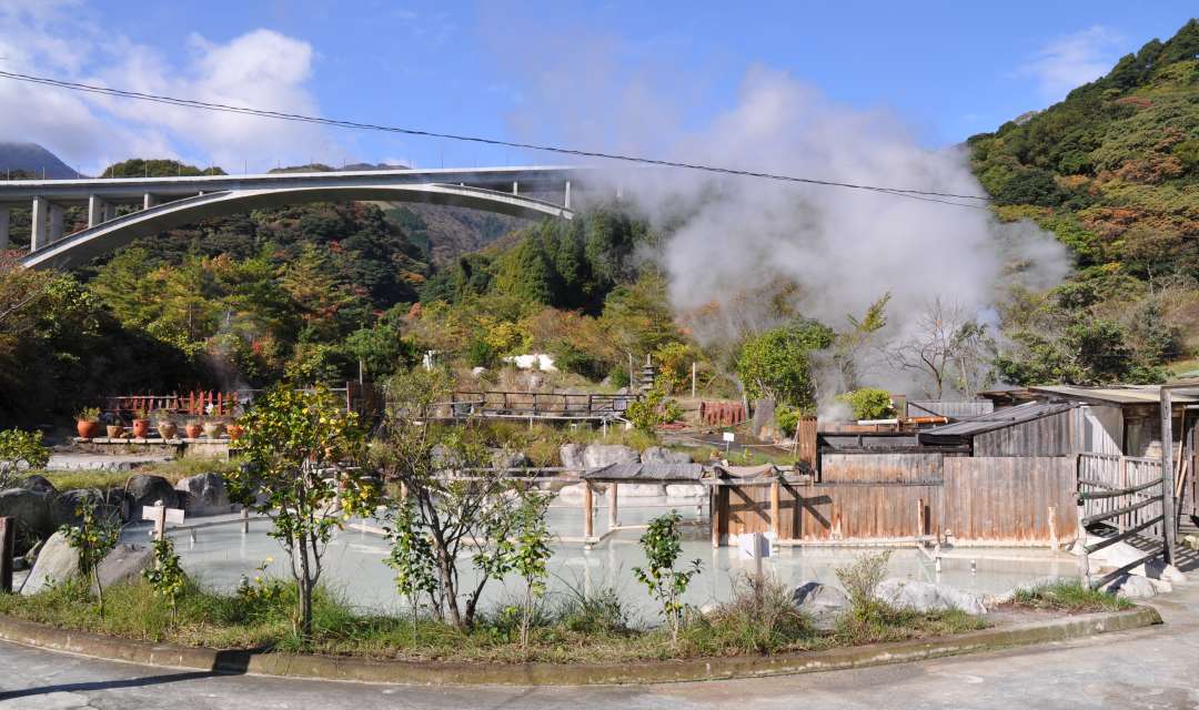 An onsen pool in the foreground and steam, trees and bridge in the background