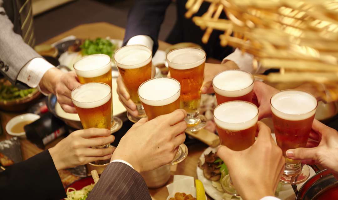 eight glasses of beer held by hands above food on a table