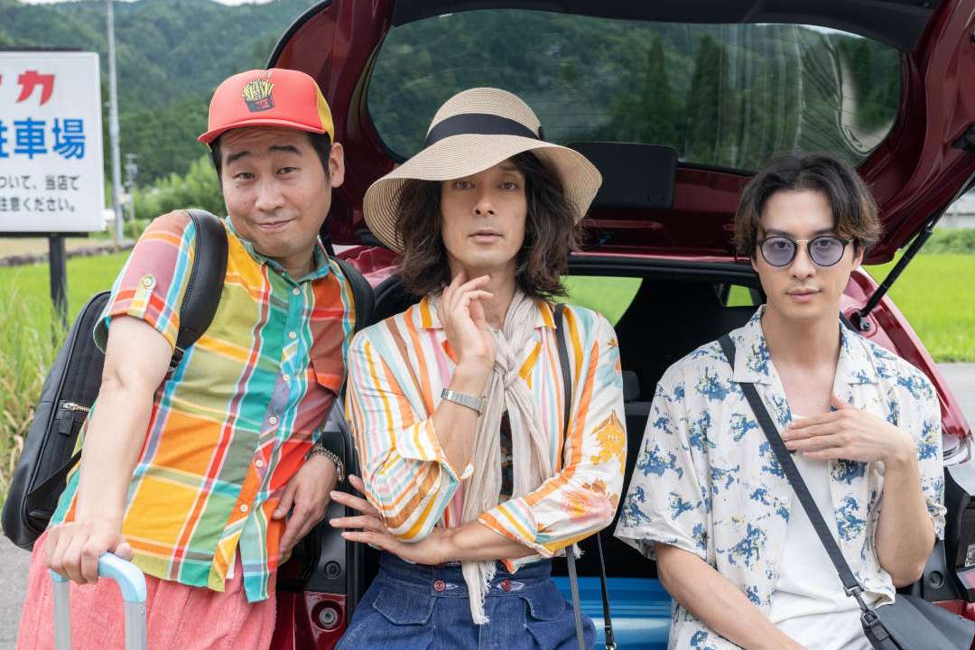 Promotional image for the film Natchan’s Little Secret, with 3 people leaning against the back of a car and posing for the camera