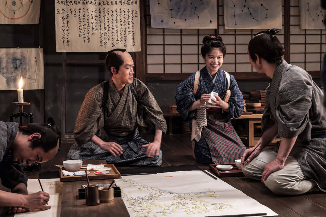 4 people wearing traditional Japanese clothes inside a traditional Japanese house, kneeling around a map of Japan