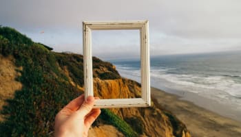 a hand holding a picture frame over a cliff with a beach and water