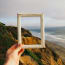 a hand holding a picture frame over a cliff with a beach and water