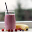 a glass of pink smoothie with straw next to a banana and berries