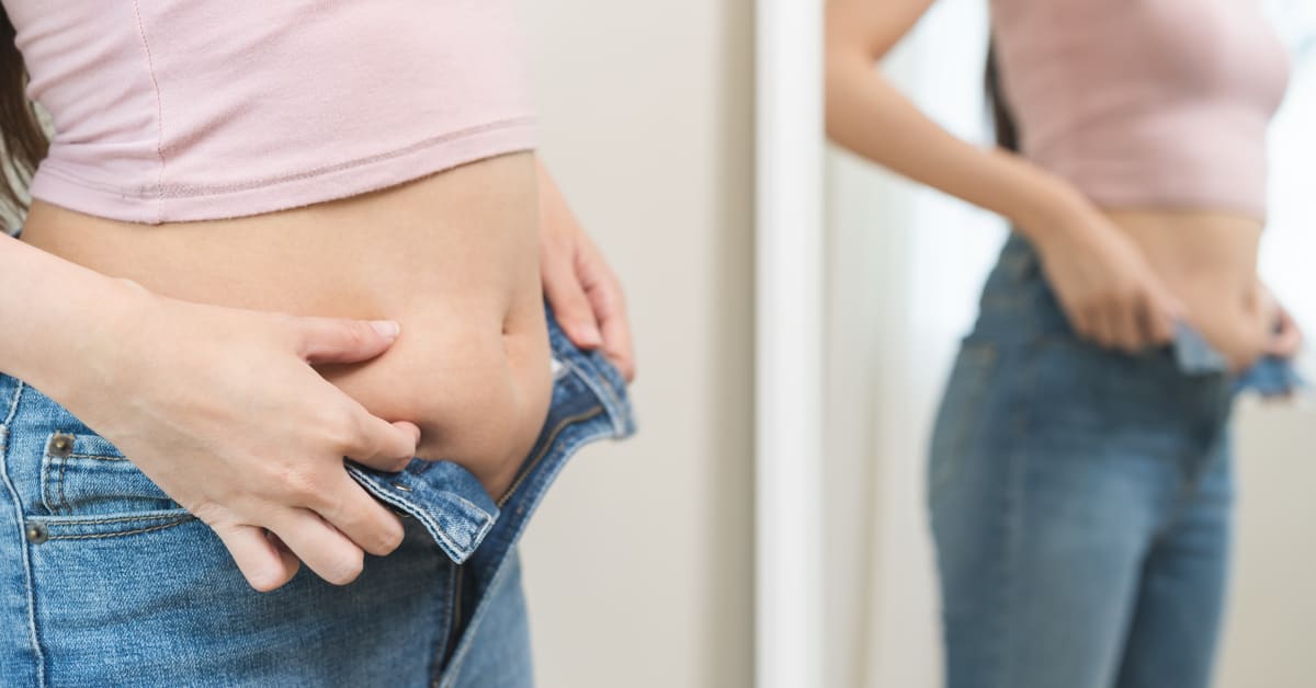image of woman pinching at stomach while trying to button up pants image of woman pinching at stomach while trying to button up pants