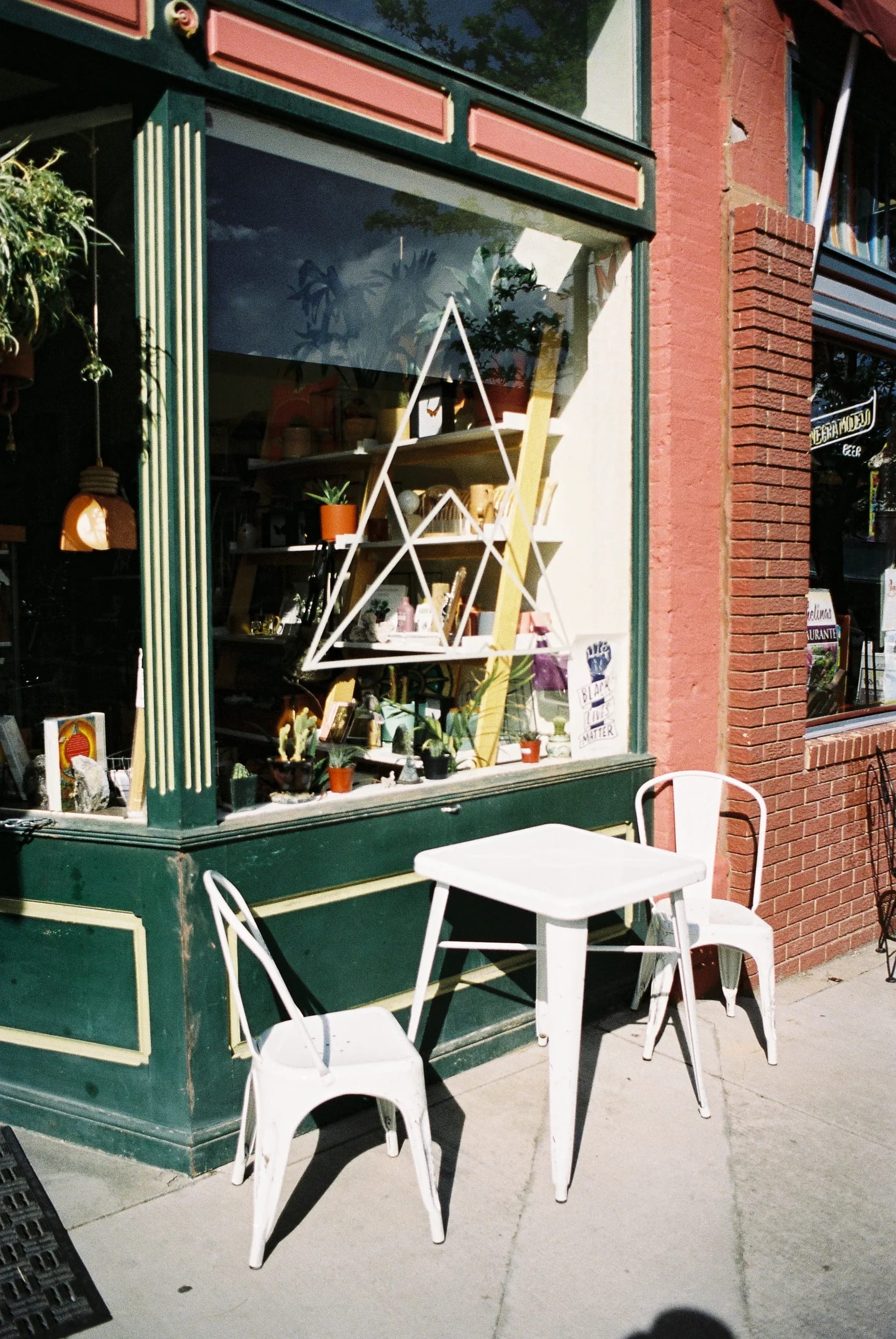 Sunlit street view of a small shop with green and red facade, display window filled with plants, and a white table with chairs on the sidewalk.