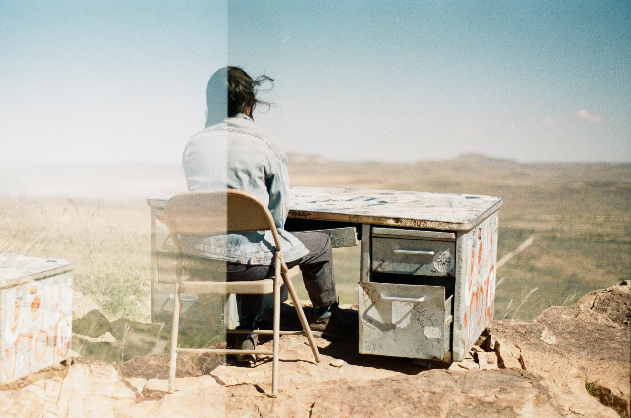 Double exposure: figure at a graffiti-tagged desk on a cliff edge, overlooking open desert.