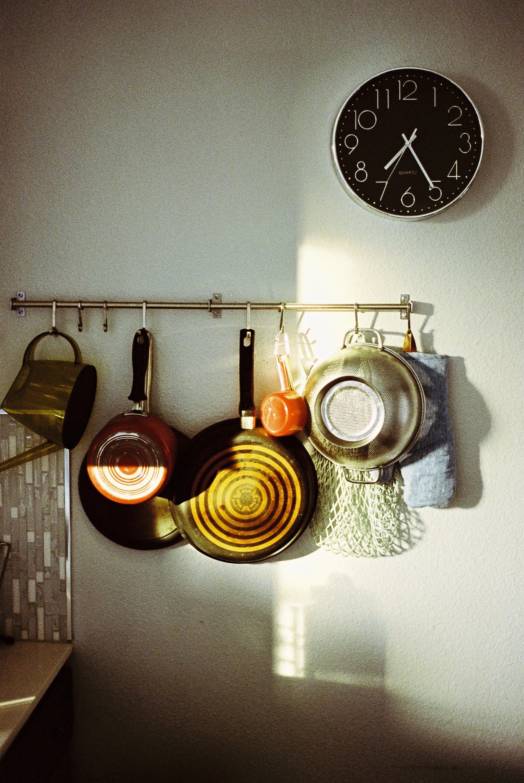 Wall-mounted black clock and rail with hanging kitchen utensils—frying pans, colander, copper pot—illuminated by a sunbeam.