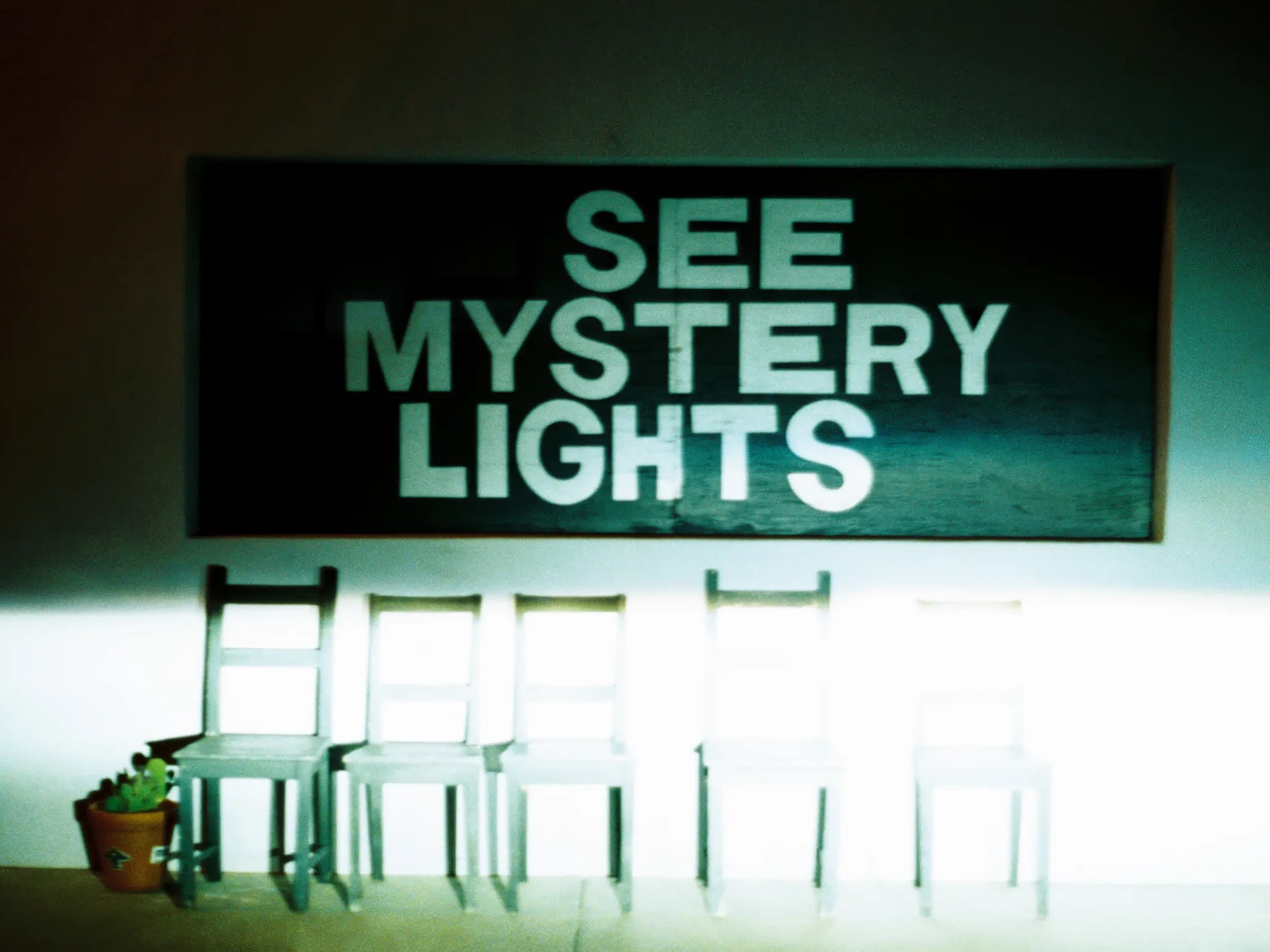 "SEE MYSTERY LIGHTS" sign above five wooden chairs with a potted cactus at one end.