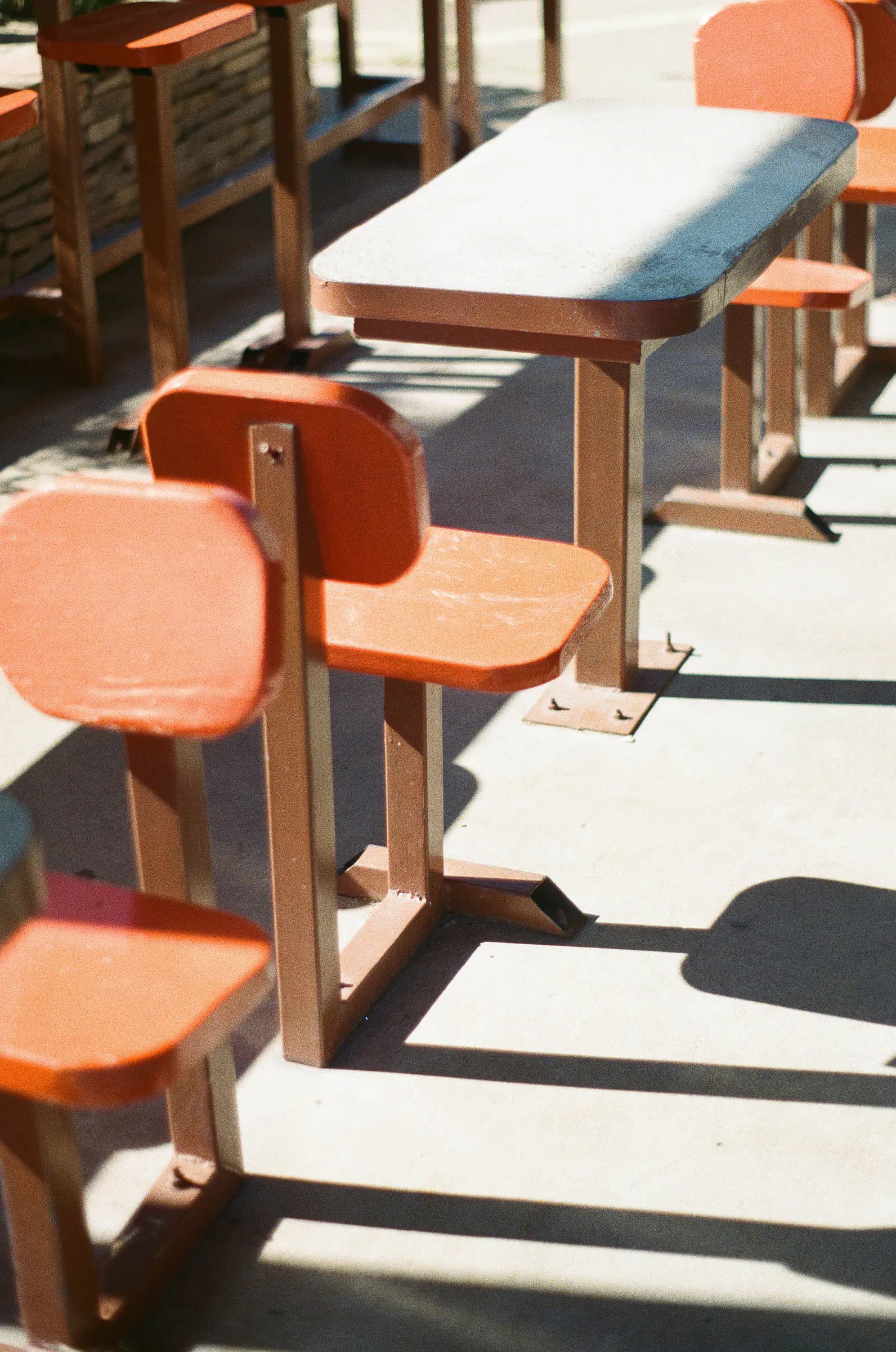 Orange-brown fixed tables and chairs on concrete, harsh shadows in bright sunlight.