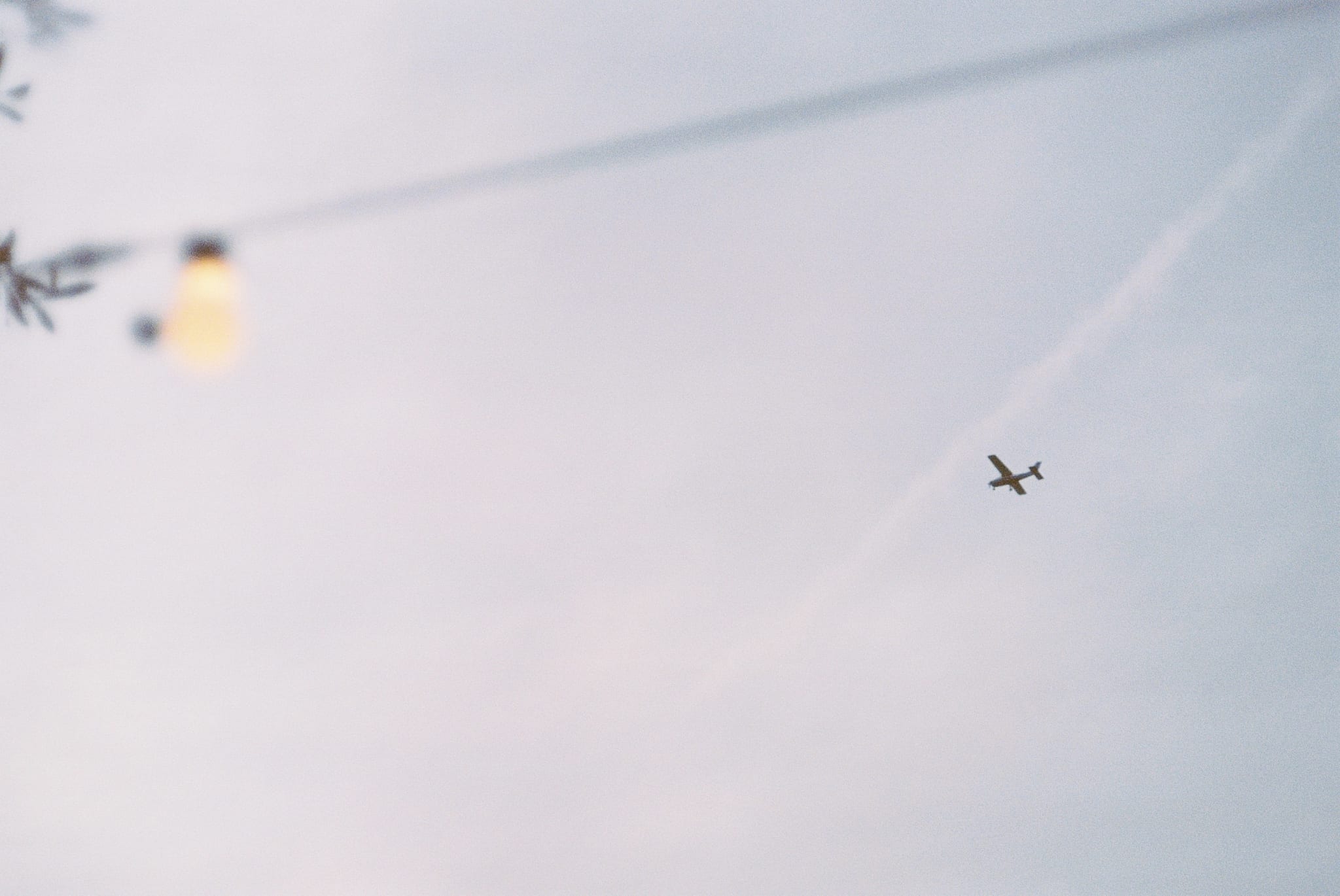 Small airplane crosses a pale cloudy sky with faint contrail. Blurry string lights and foliage frame the upper left foreground.