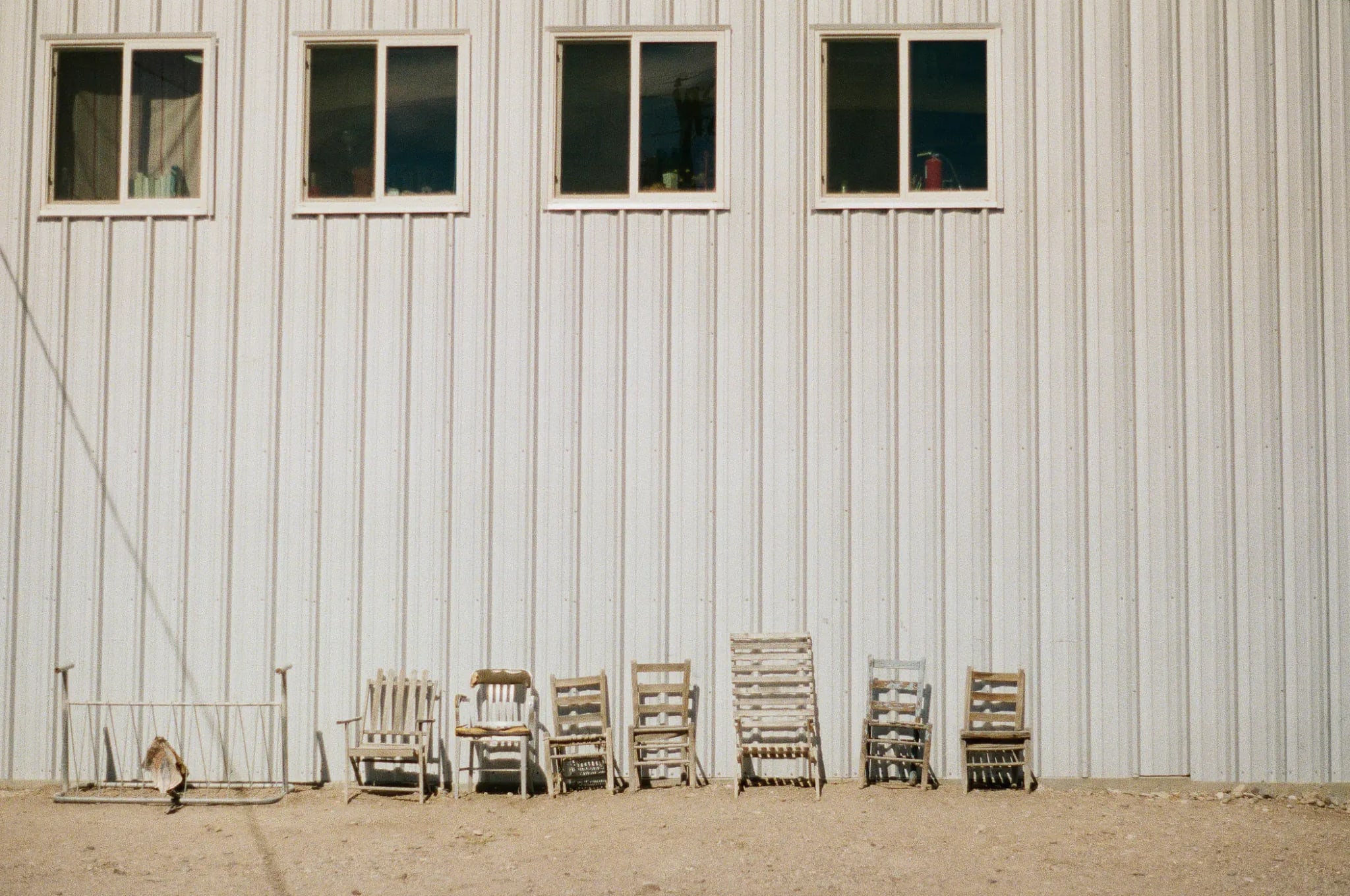Rustic wooden chairs lined up on gravel outside a white metal building under a clear blue sky.