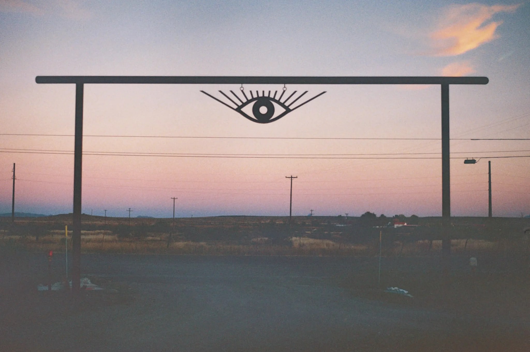 Metal archway with an all-seeing eye symbol silhouetted against a dusk sky, overlooking a sparse desert with distant utility poles.