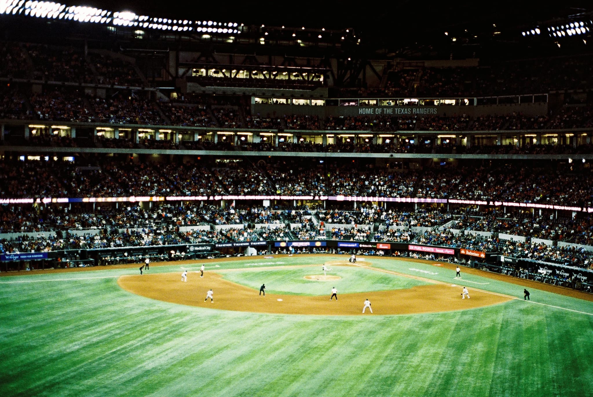 Elevated view of a crowded baseball stadium at night with players on the field, tiered seating, and a 'Home of the Texas Rangers' sign in the upper deck.