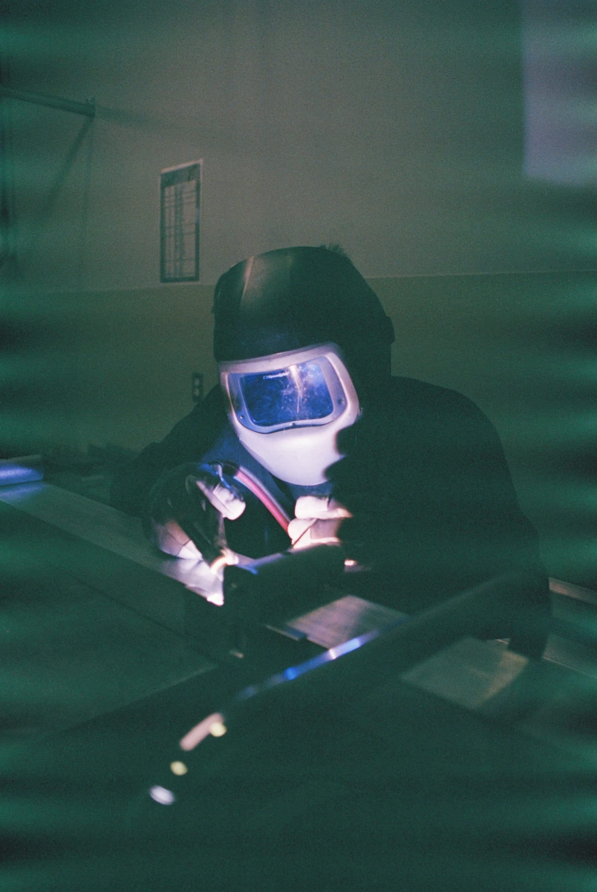 Welder in an auto-darkening helmet, shown from chest up, with bright arc light visible through the visor and green light streaks across the frame.
