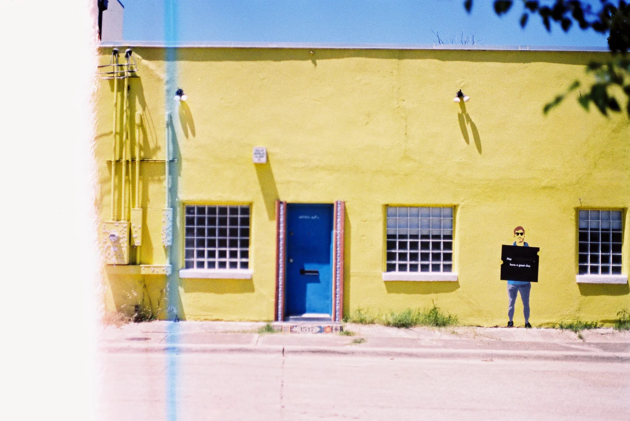 Yellow building with blue door and gridded windows. A figure holds a sign reading "Hey have a great day."