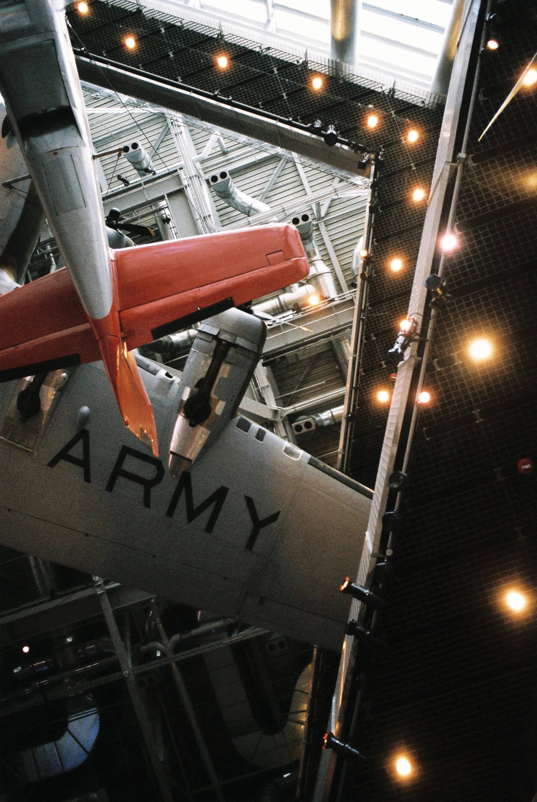 Underside view of a vintage gray U.S. Army transport aircraft with red tail, suspended in a museum hangar beneath industrial ceiling structures.