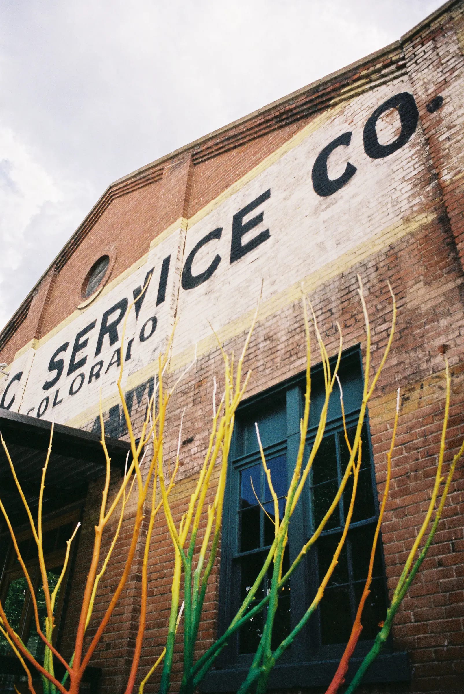 Faded "SERVICE CO. COLORADO" sign on red brick, framed by colorful painted branches.