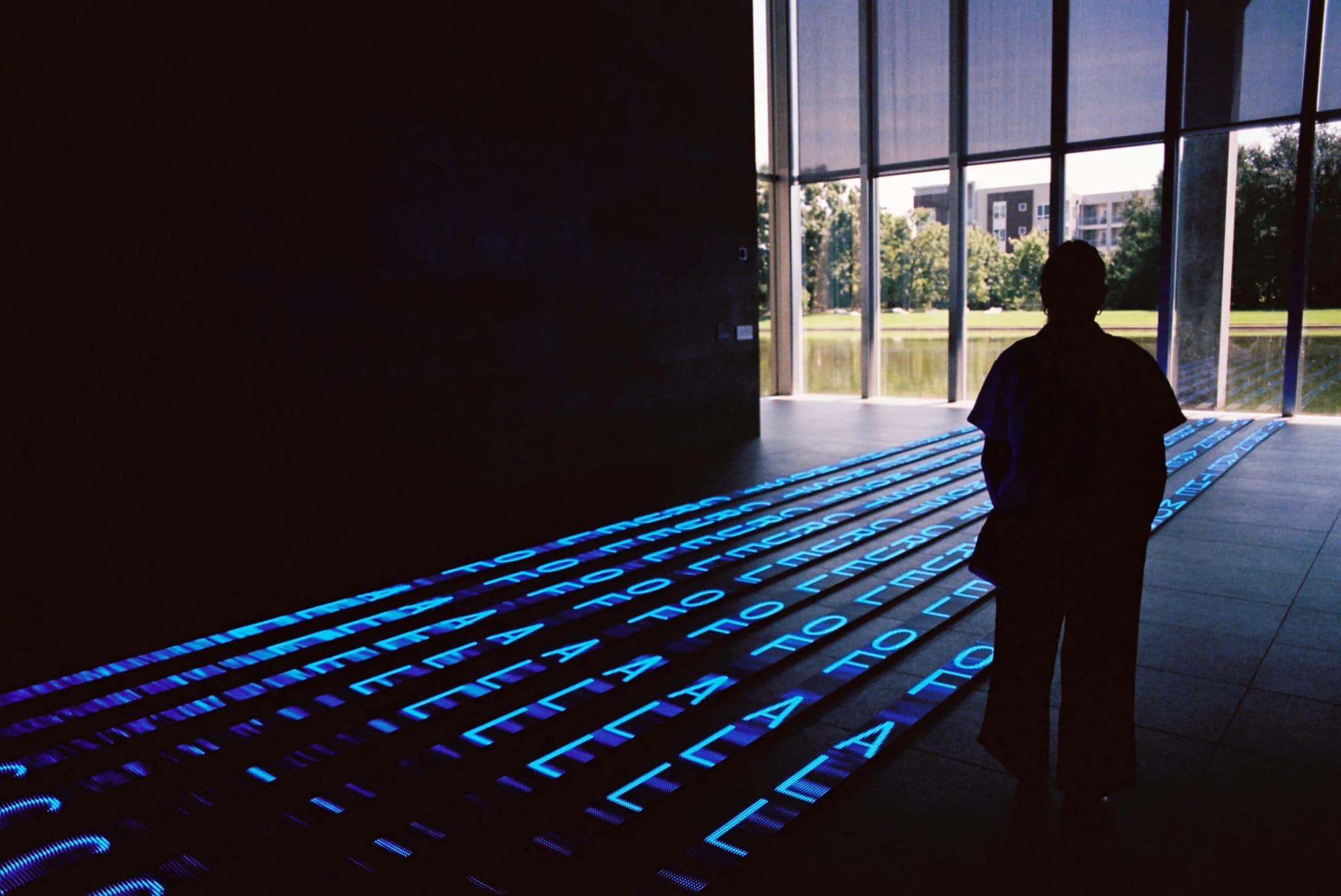 Silhouetted figure in a dim room faces large windows overlooking a pond. Glowing blue text projects onto the floor in parallel lines toward the view.