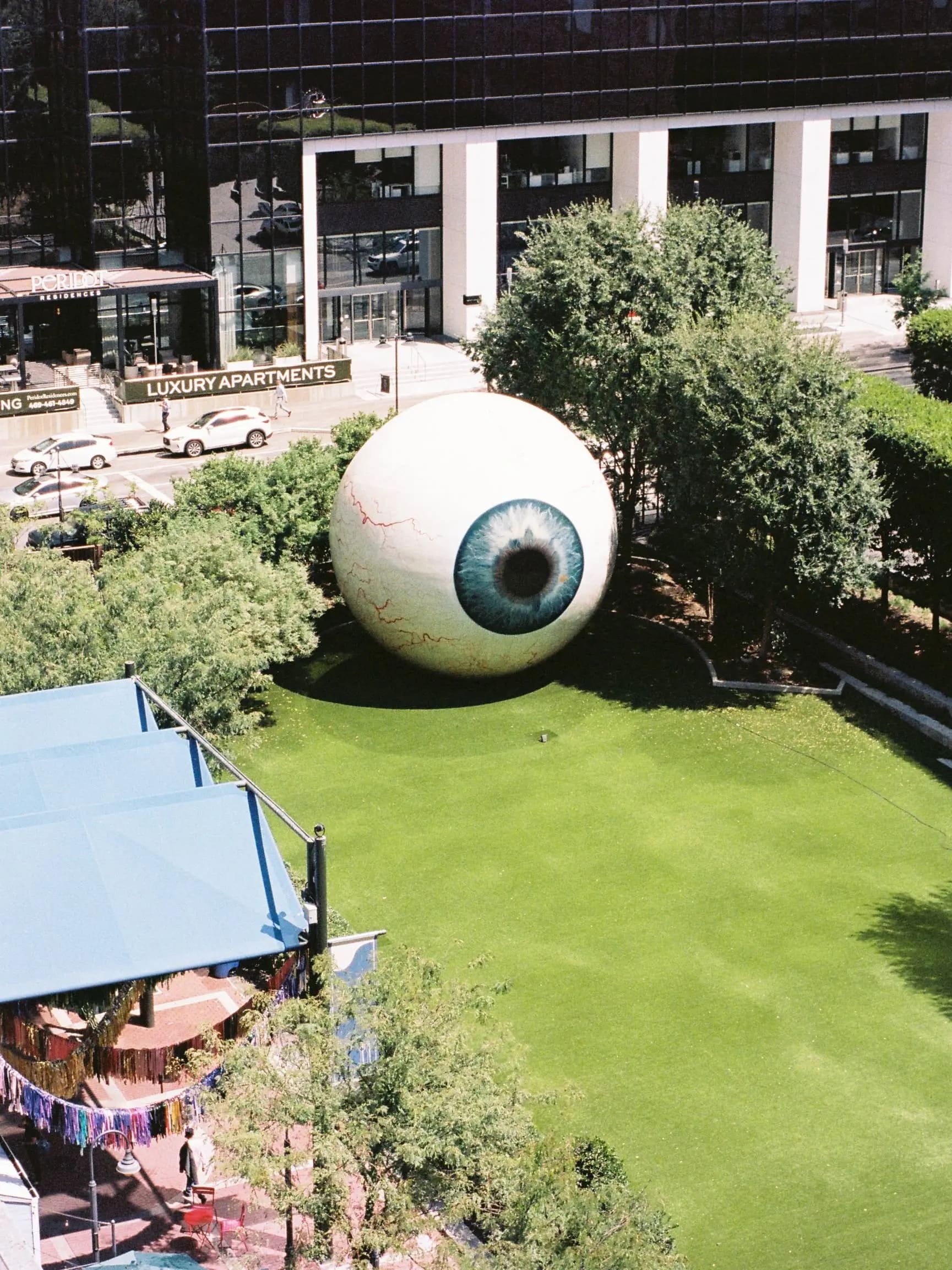 Giant eyeball sculpture on a lawn, framed by trees and a modern apartment building