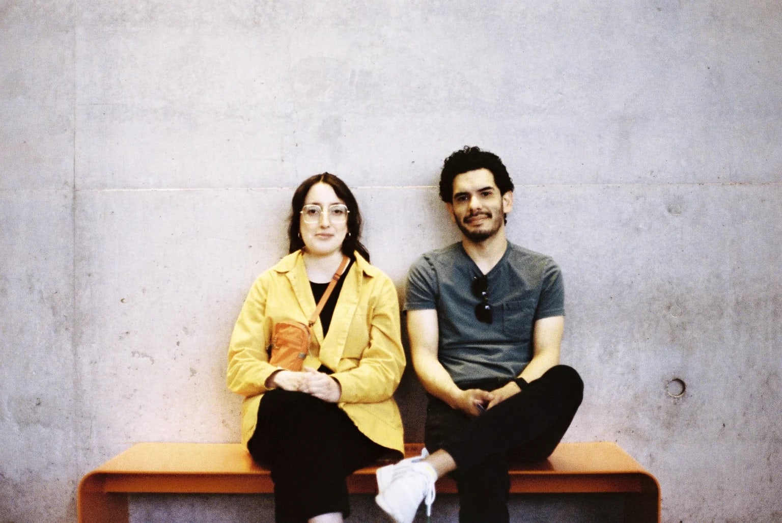 Couple on an orange bench against a concrete wall, both smiling at the camera.