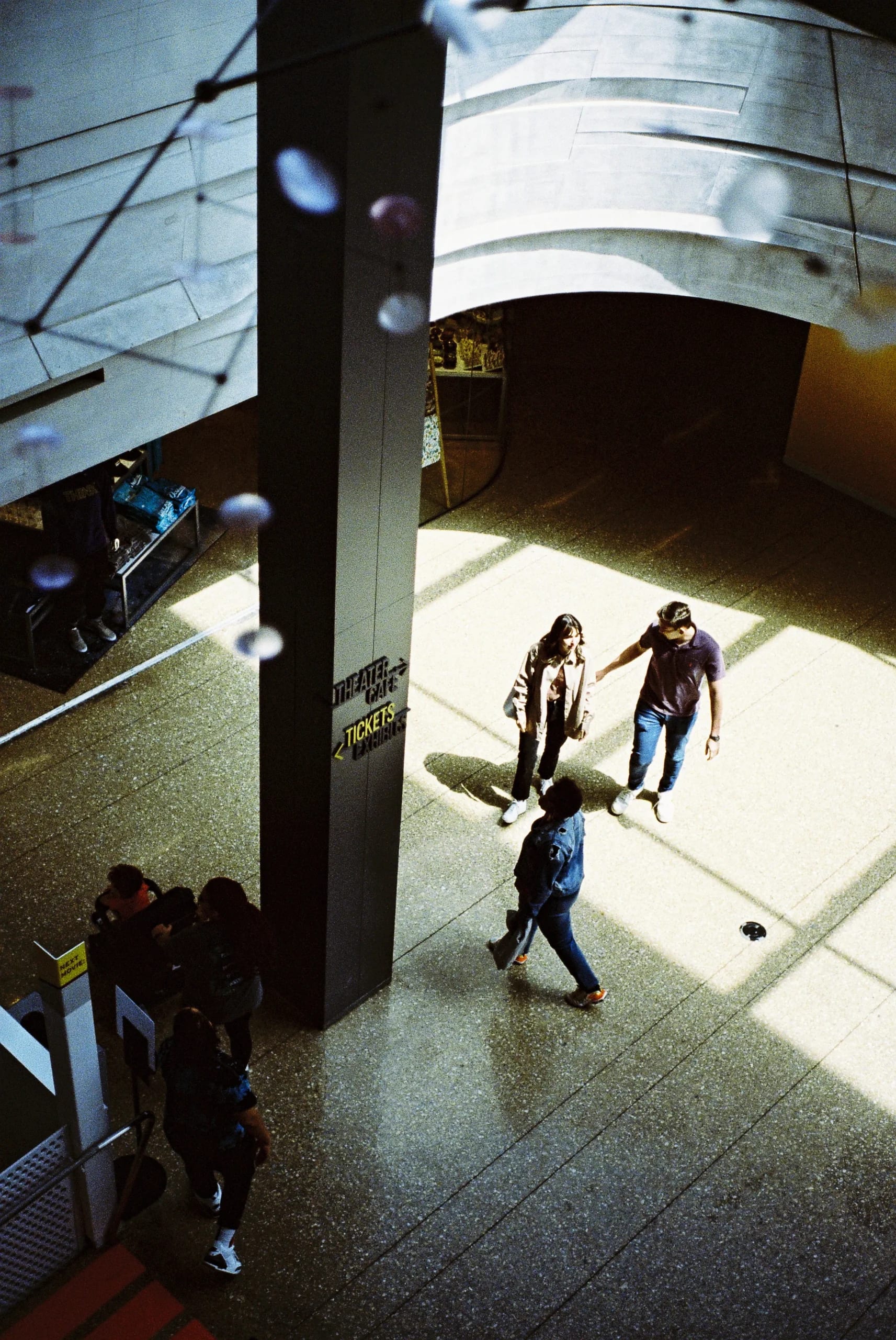 Overhead view of a museum hall with people crossing a speckled floor. A dark pillar displays 'Theater Cafe' and 'Tickets Exhibits' signs as sunlight casts geometric shadows below.