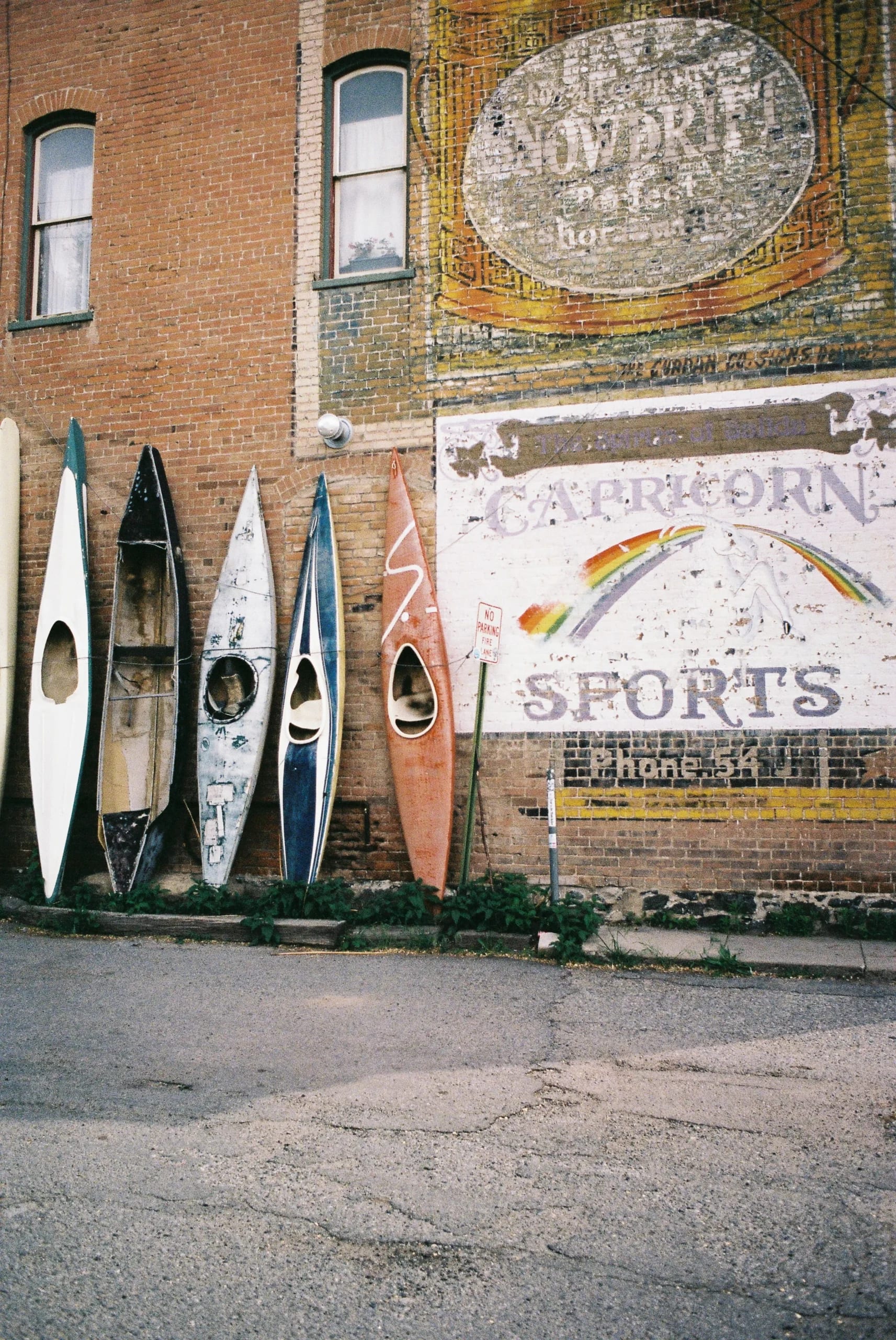 Weathered kayaks lean against a brick building with a faded 'Capricorn Sports' mural and rainbow.