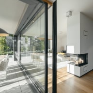 Innenansicht eines modernen Wohnbereichs mit bodentiefem Ganzglassystem von Josko, das Innen- und Außenraum verbindet. Links führt eine große Glasschiebetür zur Terrasse mit Sitzbereich, rechts steht ein weißer Kamin mit Glasfront. Der helle Raum hat Parkettboden, ein großes Sofa und wirkt lichtdurchflutet dank der großen Fensterflächen und Ausblick ins Grüne.