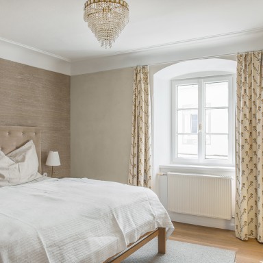 Gemütliches, helles Schlafzimmer mit weißem, klassischem Josko Fenster, einem Bett mit gepolstertem Kopfteil, cremefarbenen Vorhängen und einem gelben Sessel mit Muster.