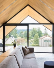 Wohnzimmer mit grauem Sofa vor einer großen Glasfront von Josko unter schräger Holzdecke, Blick auf Balkon und grüne Landschaft.