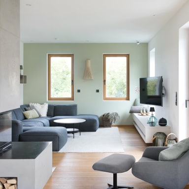 Moderner Wohnbereich mit hellgrüner Wand, grauer Eckcouch, Sessel mit Hocker und einem Kamin im Vordergrund. Zwei Holzrahmenfenster von Josko lassen Tageslicht herein, daneben steht ein Fernseher auf einem weißen Lowboard.