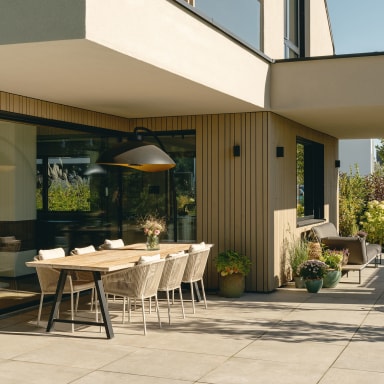 Überdachte Terrasse eines modernen Einfamilienhauses mit großen Glasflächen von Josko. Ein langer Holztisch mit sechs hellen Stühlen steht im Mittelpunkt, darüber eine große, dunkle Hängelampe. Rechts befindet sich eine gemütliche Sitznische mit grauer Outdoor-Liege und mehreren Pflanzen in Töpfen. Vertikale Holzlamellen verkleiden die Fassade, umgeben von dicht bepflanztem Garten mit Sträuchern und Blumen bei sonnigem Tageslicht.