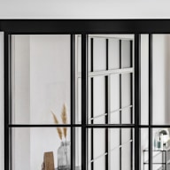 Detail einer raumhohen Loft-Schiebetür mit schwarzem Metallrahmen und Sprossen, Blick in einen hellen Wohnbereich mit minimalistischer Einrichtung.
