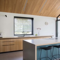 Moderne Küche mit Holzdecke, schwarzer Schrankwand, großer Kochinsel mit weißer Arbeitsplatte, zwei schwarzen Barhockern und großer Josko Glasfront.
