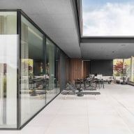 Große, überdachte Terrasse mit hellem Fliesenboden, bodentiefen Glasflächen von Josko mit Vorhängen auf der linken Seite, mehreren Sonnenliegen, Esstisch und Lounge-Bereich im Hintergrund sowie einem eingelassenen Pool auf der rechten Seite, umgeben von grüner Hügellandschaft.