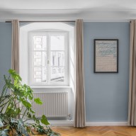 Wohnraum mit zwei weißen, klassischen Josko Fenstern, hellblauen Wänden, beigen Vorhängen, einem abstrakten Bild an der Wand und einer großen Monstera-Pflanze im Vordergrund.