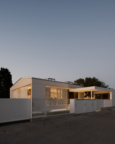 Modernes, eingeschossiges Wohnhaus mit klarer, reduzierter Architektur hinter einem weißen Zaun und Tor. Große Fensterflächen mit dekorativen, halbtransparenten Elementen geben den Blick ins warm beleuchtete Innere frei. Rechts schließt ein überdachter Außenbereich mit Flachdach an. Die Szene ist in der Dämmerung aufgenommen, die nasse Straße im Vordergrund spiegelt das Licht.
