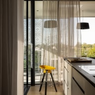 Moderner Küchenbereich mit großer Fensterfront und halbtransparenten Vorhängen, durch die warmes Tageslicht fällt. Eine helle Kücheninsel mit Arbeitsfläche steht im Vordergrund, daneben ein hoher Hocker mit gelber Sitzfläche. Dekorative Sichtschutzelemente auf dem Balkon filtern das Licht und schaffen ein ruhiges, wohnliches Ambiente.