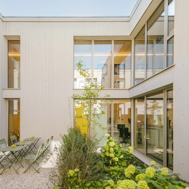 Heller Innenhof eines modernen Gebäudes mit hellen Holzfassaden und großen Glasflächen von Josko. In der Mitte stehen grüne Pflanzen, daneben ein kleiner Sitzbereich mit grünen Metallstühlen und Tischen auf hellem Kiesboden.