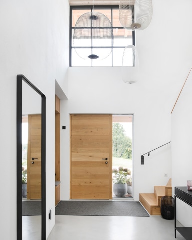 Lichtdurchfluteter Eingangsbereich mit charakterstarker Holztür von Josko und großem, schwarzem Sprossenfenster – moderne Architektur, die Natürlichkeit und Design harmonisch verbindet.