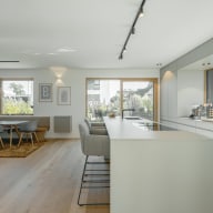 Offener Wohn- und Küchenbereich mit viel Tageslicht, großen Fenstern und Holzfußboden. Links steht ein Esstisch mit Sitzbank, Stühlen und zwei dekorativen Holzpendelleuchten darüber. Rechts befindet sich eine moderne, helle Küche mit langer Kücheninsel, flankiert von zwei gepolsterten Barhockern. Eine breite Fensterfront über der Arbeitsfläche gewährt Ausblick auf den Garten. Bodentiefe Schiebetüren verbinden den Innenraum mit der Terrasse und lassen das Licht großzügig einfallen.