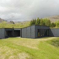Modernes Haus mit dunkler Holzfassade und großen Fenstern, eingebettet in eine grüne Landschaft vor felsigen Bergen.