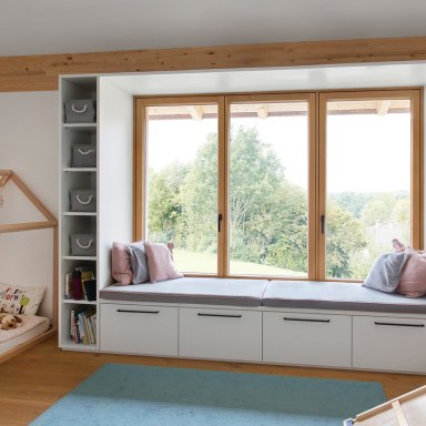 Helles Kinderzimmer mit großem Holzfenster von Josko und eingebauter Fensterbank mit Kissen. Links steht ein offenes Kinderbett aus Holz in Hausform, daneben Regale mit grauen Stoffboxen und Kinderbüchern.