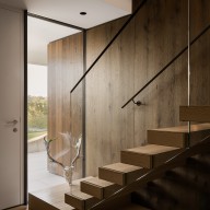 Moderner Innenbereich mit offener Holztreppe und klarer, reduzierter Gestaltung. Großzügige Glasflächen neben der weißen Haustür lassen viel Tageslicht herein und geben den Blick nach draußen frei. Die warme Holzverkleidung an Wand und Stufen schafft eine wohnliche Atmosphäre, während ein filigranes Geländer die Treppe begleitet.