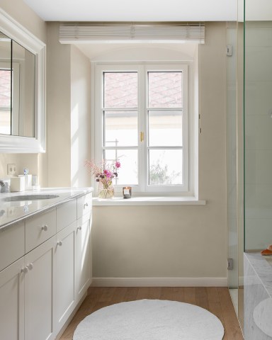 Modernes, helles Badezimmer mit weißem, klassischem Josko Fenster, einer Waschtischzeile aus weißem Holz mit Marmorabdeckung, einem runden Teppich und einer Glasdusche rechts.