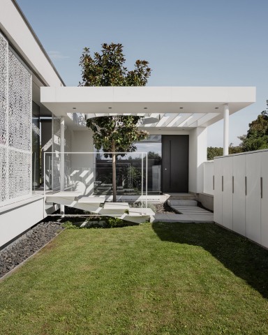 Moderner Eingangsbereich eines Einfamilienhauses mit klarer, weißer Architektur. Eine schwarze Haustür von Josko liegt leicht zurückgesetzt unter einem flachen Vordach. Eine kleine Treppe mit schwebenden Stufen und Glasgeländer führt über einen begrünten Innenhof mit Baum zum Eingang. Große Fensterflächen und dekorative, durchbrochene Fassadenelemente sorgen für Licht und Transparenz.