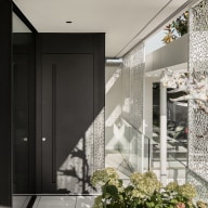 Moderner Eingangsbereich mit dunkler, minimalistischer Haustür und seitlicher Glasfläche. Rechts verlaufen große Glasflächen mit dekorativen, filigranen Sichtschutzelementen, die ein lebendiges Licht- und Schattenspiel erzeugen. Ein Glasgeländer begleitet den Zugang, im Vordergrund sorgen Pflanzen für eine natürliche Note. Sonnenlicht durchflutet den offenen, architektonisch klar gestalteten Bereich.