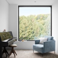 Heller, minimalistischer Raum mit großem quadratischem Fenster von Josko mit schwarzem Rahmen, davor ein hellblauer Sessel mit Kissen, links ein schwarzes Klavier mit rundem Hocker, durch das Fenster Blick auf dichten grünen Wald.