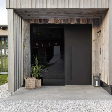 Moderne Hauseingangssituation mit dunkler Josko Haustür in glatter Oberfläche, umrahmt von einer Holzfassade. Vor dem Eingang stehen dekorative Pflanzen und Laternen auf hellem Kiesboden.