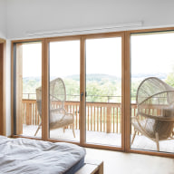 Lichtdurchflutetes Schlafzimmer mit großzügigen Holz-Alu-Fenstern und Zugang zum Balkon – natürliche Materialien und weiter Ausblick schaffen Ruhe und Geborgenheit.